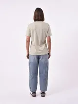 Remera de manga corta color beige claro con cuello redondo. Presenta un estampado gráfico central con un plato de pasta rodeado por un borde azul y blanco, sobre un fondo de franjas verticales rojas y blancas. El estampado incluye texto en cursiva en tonos turquesa y rojo.