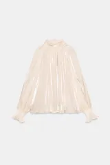Camisa satinada color beige claro, de cuello alto fruncido, mangas largas abullonadas con puños elásticos y volados, y cierre posterior con botones.