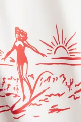 Remera de punto de algodón color blanco crudo, corte oversized con hombros caídos y cuello redondo. Estampado frontal en color rojo con una ilustración de palmeras, montañas, una surfista y el texto "Where the mountains meet the sea Santa Barbara, Calif.".