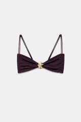 Top de bikini bordó, con escote corazón, tirantes finos dobles y frunces en los laterales. Presenta una aplicación de pieza metálica dorada con forma de sol en el centro.