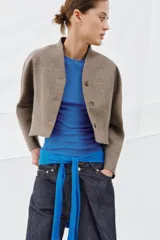 Chaqueta corta de corte estructurado confeccionada en tejido con mezcla de lana. Presenta cuello en pico, cierre frontal con botones y mangas largas. El diseño incluye bolsillos delanteros y un forro interior a contraste.