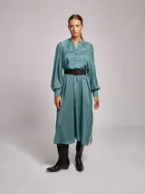 Vestido midi de satén con estampado geométrico en tonos marrones y naranjas. Presenta cuello mao, mangas largas abullonadas con puños ajustados y un cinturón a tono con hebilla metálica que marca la cintura.