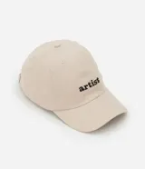 Gorra de sarga color beige con visera curva y bordado frontal con la palabra "artist" en negro. Ajuste trasero con hebilla metálica.
