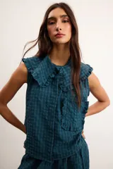 Blusa de algodón con patrón a cuadros vichy en tonos verde oscuro y azul, sin mangas, con cuello amplio decorado con volados, botones delanteros y un bolsillo frontal aplicado con ribete de volados.