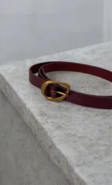 Cinto fino de cuero color bordó con hebilla metálica dorada de diseño redondeado.