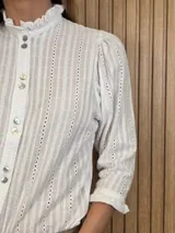 Camisa de algodón color blanco con rayas verticales rosadas, cuello alto con volados, mangas tres cuartos abullonadas con puños elastizados con volados y cierre frontal de botones oscuros.