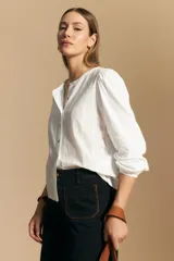 Camisa de algodón blanca con delicadas líneas bordadas verticales y textura calada. Presenta escote redondo con cierre frontal de botones, mangas largas con leve volumen en los hombros y puños elásticos.