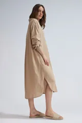 Camisa larga de lino color beige, de corte oversize, con mangas largas, cuello camisero y cierre frontal de botones. Presenta dobladillo curvo con aberturas laterales.