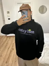 Canguro negro con capucha y cordones ajustables. Estampado frontal con la frase "estoy bien" en letras lilas y una flor verde.
