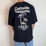 Remera negra de corte oversize con estampado gráfico en la espalda que presenta hongos en tonos violeta y verde, junto con la frase 'Curbside Treasures' y texto descriptivo.