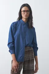Camisa de jean en tencel con silueta amplia, hombros caídos, cuello clásico y cierre frontal con botones. Presenta ruedo curvo con tajos laterales y pliegue en la espalda.