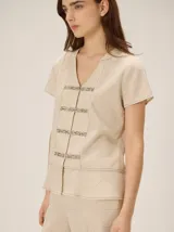 Camisa de manga corta color beige con estampado a cuadros, cuello en V y detalles de galones decorativos estilo oriental en la parte frontal.