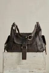 Cartera hobo de cuero marrón con efecto desgastado, solapa frontal con ojales metálicos y tira con hebilla. Cuenta con bolsillos laterales con solapa y herrajes metálicos plateados.