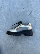 Zapato mocasín plateado con cordones negros, plataforma track negra y detalles en cuero color crema.