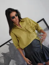 Camisa de popelina color verde oliva, de corte holgado, con cuello clásico, cierre frontal de botones, bolsillo en el pecho con detalle de rosetón a tono y diseño asimétrico más largo en la parte trasera.