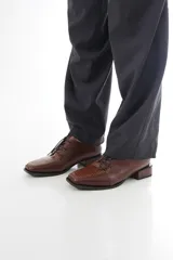 Zapato de cuero color marrón, acordonado, con puntera cuadrada y detalle texturizado en el empeine, taco bajo y acabado brillante.