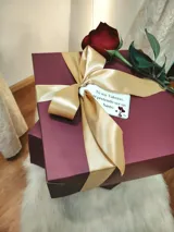 Caja de regalo artesanal color bordó con moño dorado, diseñada como experiencia de lujo para presentar un conjunto de lencería. Incluye elementos de regalo adicionales como perfume, rosa natural y dulces.