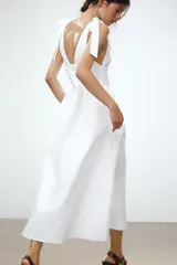 Vestido midi blanco de algodón, con escote pico pronunciado y tirantes anchos ajustables con lazada. Presenta elástico en la cintura y falda con pliegues.