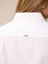 Camisa de corte clásico y calce entallado, color blanco, con cuello camisero, cierre frontal de botones y mangas largas con puños abotonados. Presenta un pequeño bordado distintivo en el pecho.