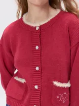 Cardigan de algodón grueso color rojo, con cuello redondo, cierre frontal de botones grandes y bolsillos cuadrados en el frente. Presenta detalles de pelo sintético en el cuello y los bolsillos, y logo bordado en uno de los bolsillos.