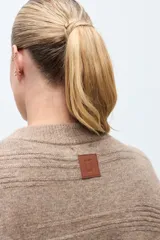 Capa tejida de punto en color beige, con diseño abierto y cierre frontal mediante broche metálico con detalles en cuero sintético negro. Presenta mangas cortas y textura suave.