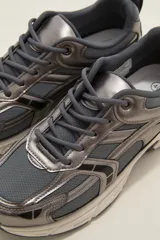 Zapatilla deportiva de estilo urbano en color blanco con detalles metalizados plateados y acentos en negro. Presenta una combinación de malla transpirable y paneles sintéticos, con suela robusta y cordones al tono.