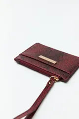 Billetera rectangular color bordó con estampado de piel de serpiente, cierre superior con cremallera, bolsillo frontal y correa de muñeca.