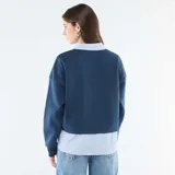 Buzo azul de cuello en V con estampa frontal, que incorpora una camisa de rayas verticales celeste y blanca debajo, creando un efecto de capas.