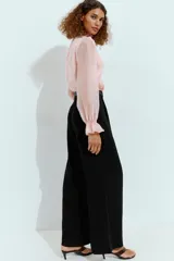 Mono largo con parte superior de blusa rosa de manga larga, cuello alto fruncido y puños con volantes. La parte inferior es un pantalón negro de pernera ancha con bolsillos delanteros y un cinturón negro con hebilla dorada.