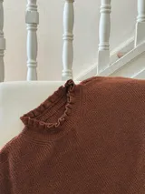 Buzo de punto oversize color rojo, con cuello alto y puños con terminaciones en volados. Presenta detalles en contraste color amarillo en el cuello y los puños, con un tejido de acabado ligeramente brillante.