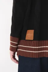 Buzo de tejido de punto con diseño de trenzas, color crema con detalles en marrón en cuello, puños y ruedo. Presenta líneas finas en contraste y pequeñas costuras decorativas en forma de cruz.