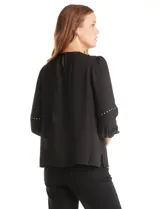 Blusa negra de gasa con cuello a la base fruncido, mangas largas amplias con puños elastizados y apliques de cinta con tachas en la parte delantera y antebrazos. Tiene una abertura central en la espalda con botón.