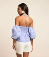Blusa de tricolina color celeste, con diseño hombro a hombro, escote halter, fruncido en el pecho con lazo ajustable, y mangas abullonadas cortas.
