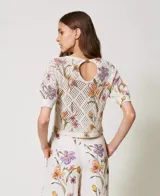 Top de tejido calado color crema con estampado floral en tonos lila, naranja y verde. Tiene cuello redondo y mangas cortas.