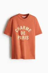 Remera de manga corta color terracota, con cuello redondo y estampado frontal en color crema que dice "Charme de Paris 1998".