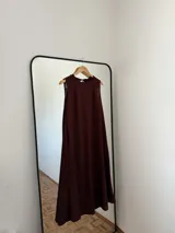 Vestido largo sin mangas, color marrón oscuro, con escote redondo y corte amplio. Presenta bolsillos laterales.
