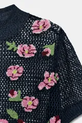 Top de punto calado color azul marino, con cuello subido y manga corta. Presenta un diseño con flores bordadas en tonos rosados y verdes en la parte delantera.