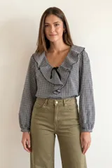 Blusa de manga larga con estampado de cuadros vichy en blanco y negro. Presenta un cuello con volados amplios y un detalle de lazo negro en el centro del escote.