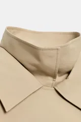 Trench corto oversize color beige con cuello subido de efecto doble, cierre cruzado con botones oscuros y bolsillos de vivo delanteros.