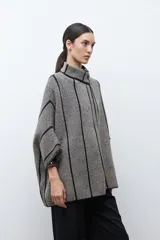 Capa de tejido de punto en color beige y negro con patrón de espiga, cuello alto y cierre de botón. Presenta vivos negros en contraste que delinean el diseño.