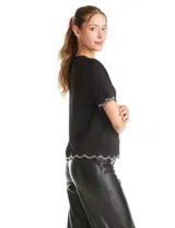 Blusa negra de crepe con cuello redondo y mangas cortas. Presenta terminaciones onduladas en mangas y ruedo, decoradas con una cinta de strass brillante. Cuenta con cierre invisible en la espalda.