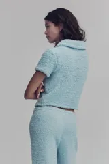 Saco corto de manga corta color celeste confeccionado en sherpa suave. Presenta cuello camisero, cierre frontal de botones metálicos dorados, bolsillos de parche en el pecho con solapa y botones, y diseño de bajo recto.