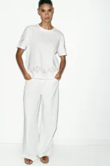 Camiseta de algodón de corte regular, color blanco, con cuello redondo y manga corta. Presenta detalles de bordados florales y ornamentales en color rosado en el ruedo y en las mangas.