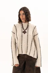 Buzo de tejido de punto color crudo con diseño de trenzas y costuras decorativas en color negro que recorren la prenda en forma de espiral. Presenta cuello redondo y mangas largas con terminación acampanada.