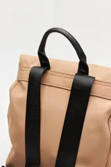 Mochila urbana de nylon color beige, con solapa superior, bolsillo frontal con cierre y correas decorativas con hebillas metálicas. Incluye manija superior y correas ajustables.