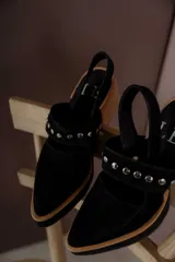 Zapatos tipo zueco de gamuza negra con punta fina y tira sobre el empeine decorada con tachas metálicas plateadas. Posee taco cuadrado de madera y suela con plataforma.