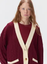 Cardigan de tejido de punto con diseño de ochos, corte oversize y mangas largas con puños acanalados. Presenta un escote en V con solapas en contraste, cierre frontal con botones y dos bolsillos delanteros con bordes en contraste. El diseño incluye detalles de rayas en los puños y en el ruedo.