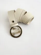 Cinto de cuero color crudo con forro de cuero y sistema de broche suizo intercambiable. Presenta una hebilla circular de guampa con borde metálico labrado en tono bronce.