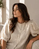 Blusa de textura sutil con acabado brillante, color beige, de corte estructural y manga corta abullonada con tablas. Presenta cuello redondo, cartera frontal con botones de nácar y largo extendido.