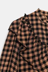Blusa de manga larga con estampado de cuadros vichy en tonos marrón y negro. Presenta cuello redondo, cierre frontal con botones y detalle de volantes en el pecho.